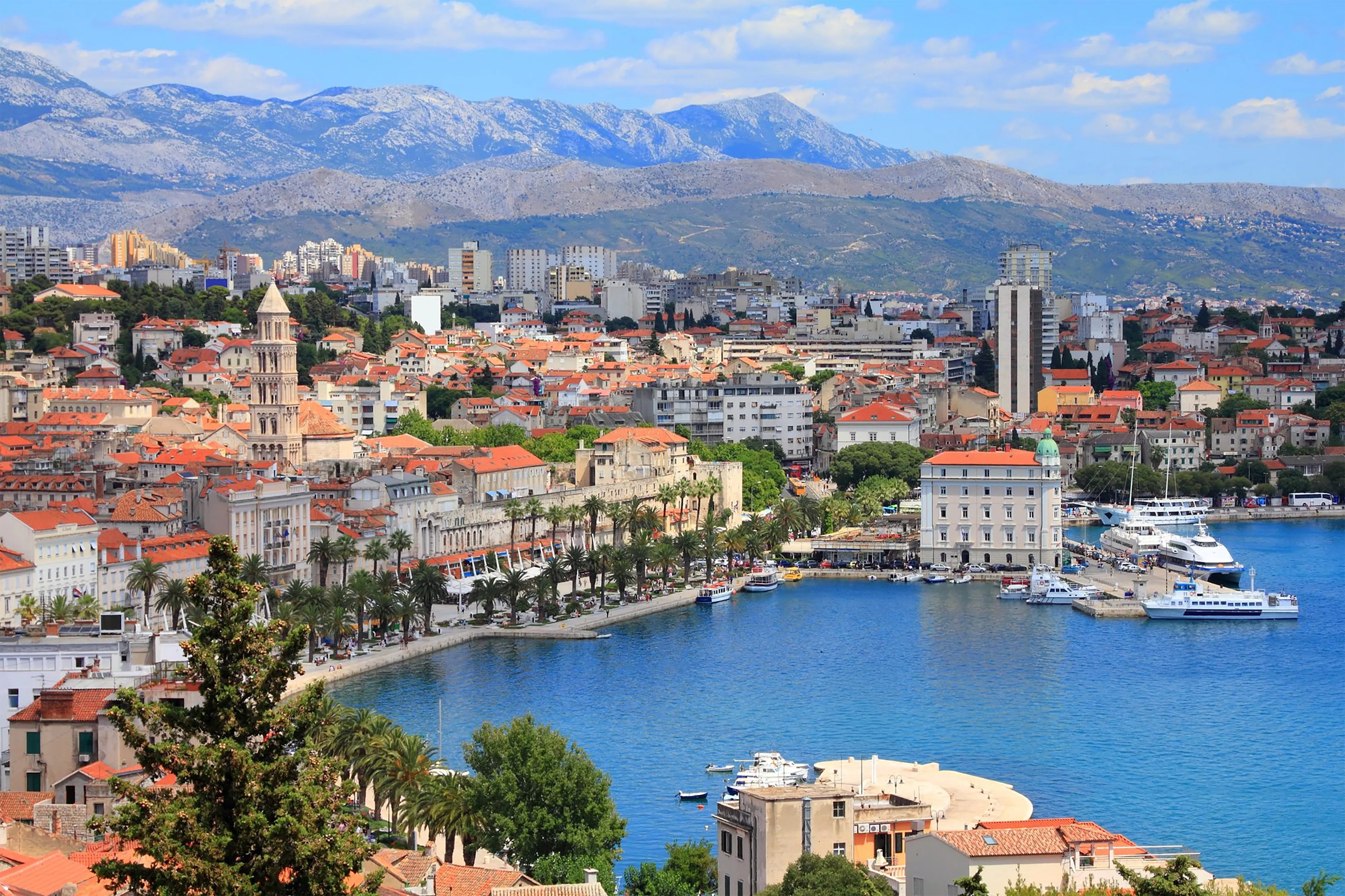 Panoramic view of Split harbor with red roofed historic buildings, waterfront promenade, boats, and mountains in the background.