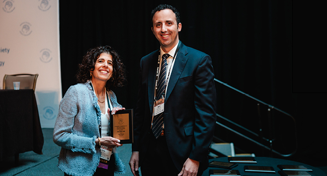 Woman with curly hair holding a plaque stands beside a man in a suit